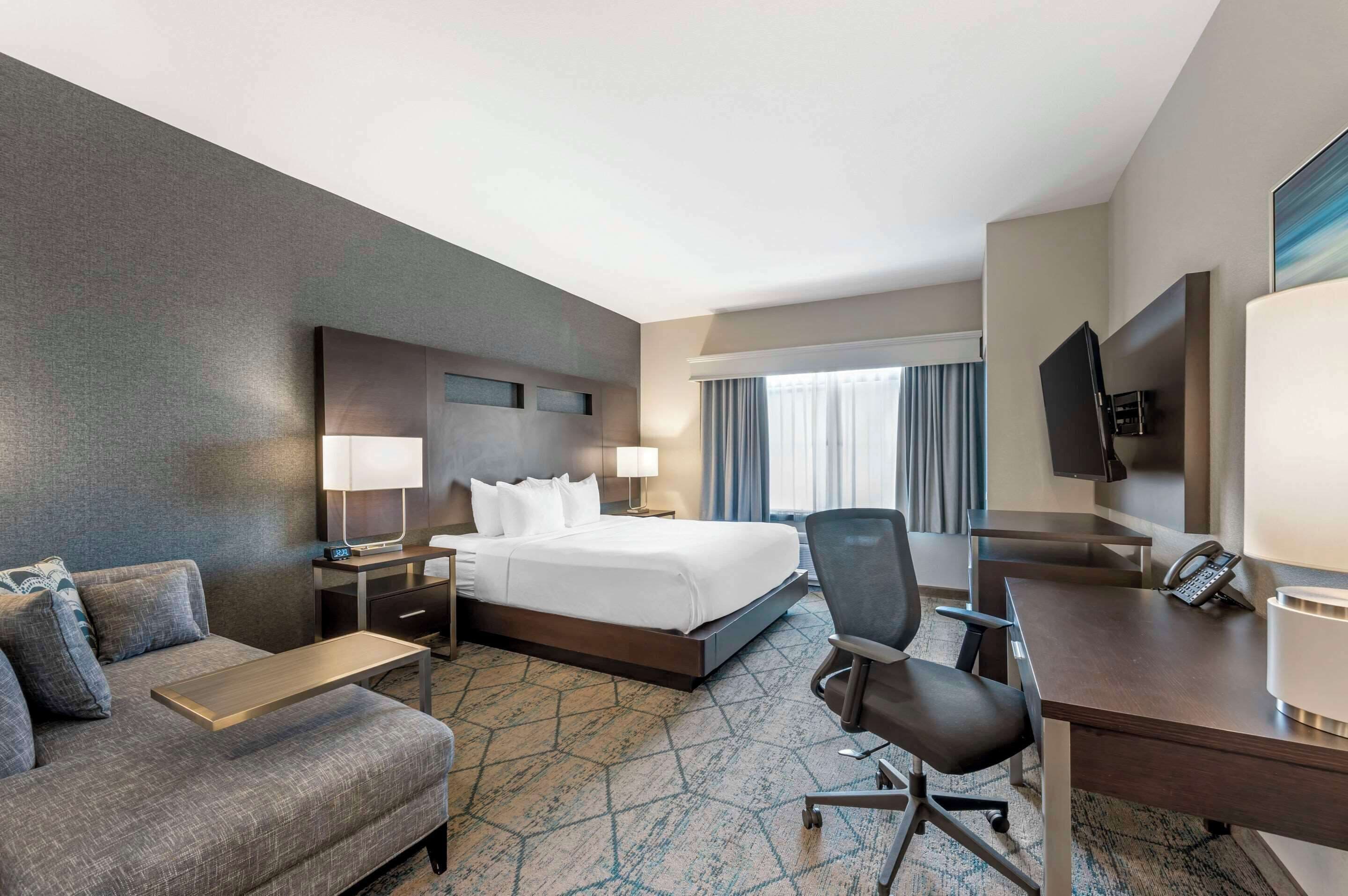A hotel room with a bed, a desk, a chair, a sofa, a TV, and a window with curtains.