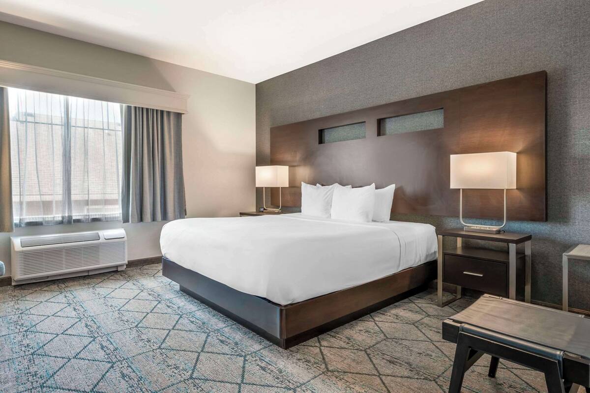 Una habitación de hotel con una cama grande, dos mesitas de noche con lámparas, una ventana con cortinas, un televisor montado en la pared y una alfombra estampada.