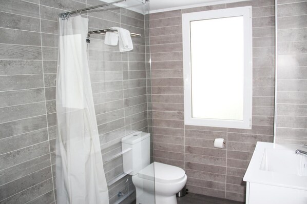 A modern bathroom with grey tiled walls, a white toilet, a shower with a white curtain, and a window providing natural light.