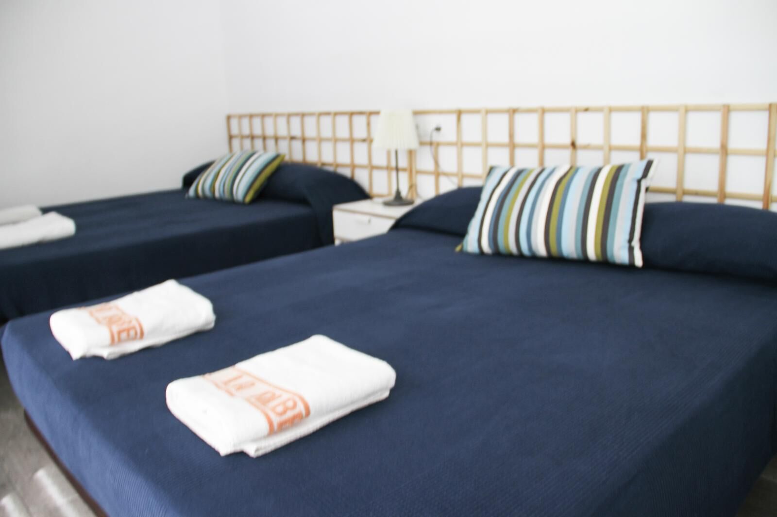 Habitación de hotel con dos camas, cada una con una colcha azul oscuro y almohadas a rayas. Hay toallas blancas con texto naranja sobre las camas.