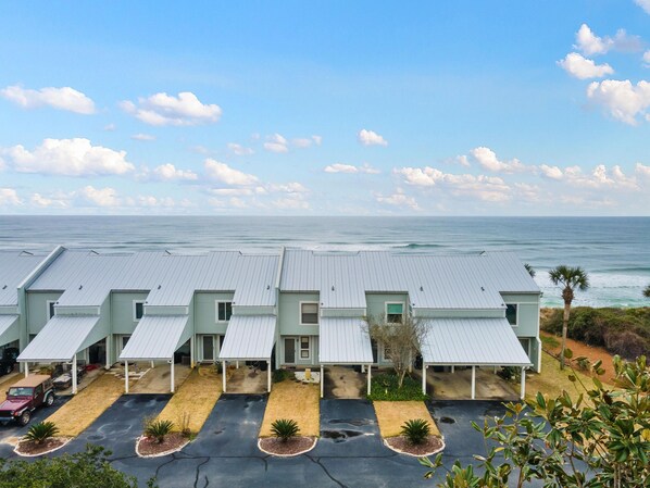 A row of light green buildings with grey roofs, a parking area with a few cars, and a view of the ocean.