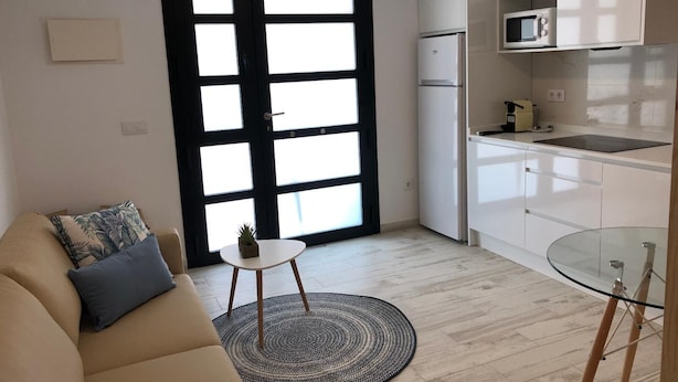 A modern living room with a sofa, a small round table, and a glass coffee table. A black-framed door with white panels is visible, leading to a kitchen area with white cabinets, a microwave, and a refrigerator. - Loft 20 meters from the sea D13 (Gandia)