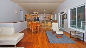 A spacious living room with a sofa, a dining area with chairs, a kitchen in the background, and large windows. -  ORCA HOUSE SPECTACULAR VIEW!! KACHEMAK DRIVE (Homer)