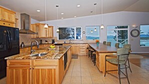 A spacious kitchen with wooden cabinets, a large island, and a view of the outdoors through large windows. -  ORCA HOUSE SPECTACULAR VIEW!! KACHEMAK DRIVE (Homer)