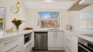 Cocina moderna con armarios blancos, lavavajillas de acero inoxidable y ventana con vista a árboles.