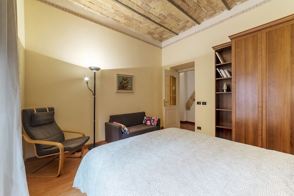 A bedroom with a bed, a chair, a sofa, a floor lamp, and a wooden wardrobe.