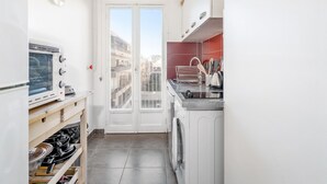 A compact kitchen with a built-in oven, a washing machine, and a window overlooking a residential area. - Appartement Corail - Welkeys (Antibes)
