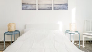 A bedroom with a bed, two nightstands, a chair, and three wall-mounted paintings. - Appartement Corail - Welkeys (Antibes)