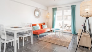 A modern living room with a white dining table, orange sofa, and a large window with teal curtains. - Appartement Corail - Welkeys (Antibes)