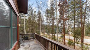 A wooden deck with a table and chairs, overlooking a forested area with tall trees. - Trapper's Crossing #8761 by Summit County Mountain Retreats (Keystone)