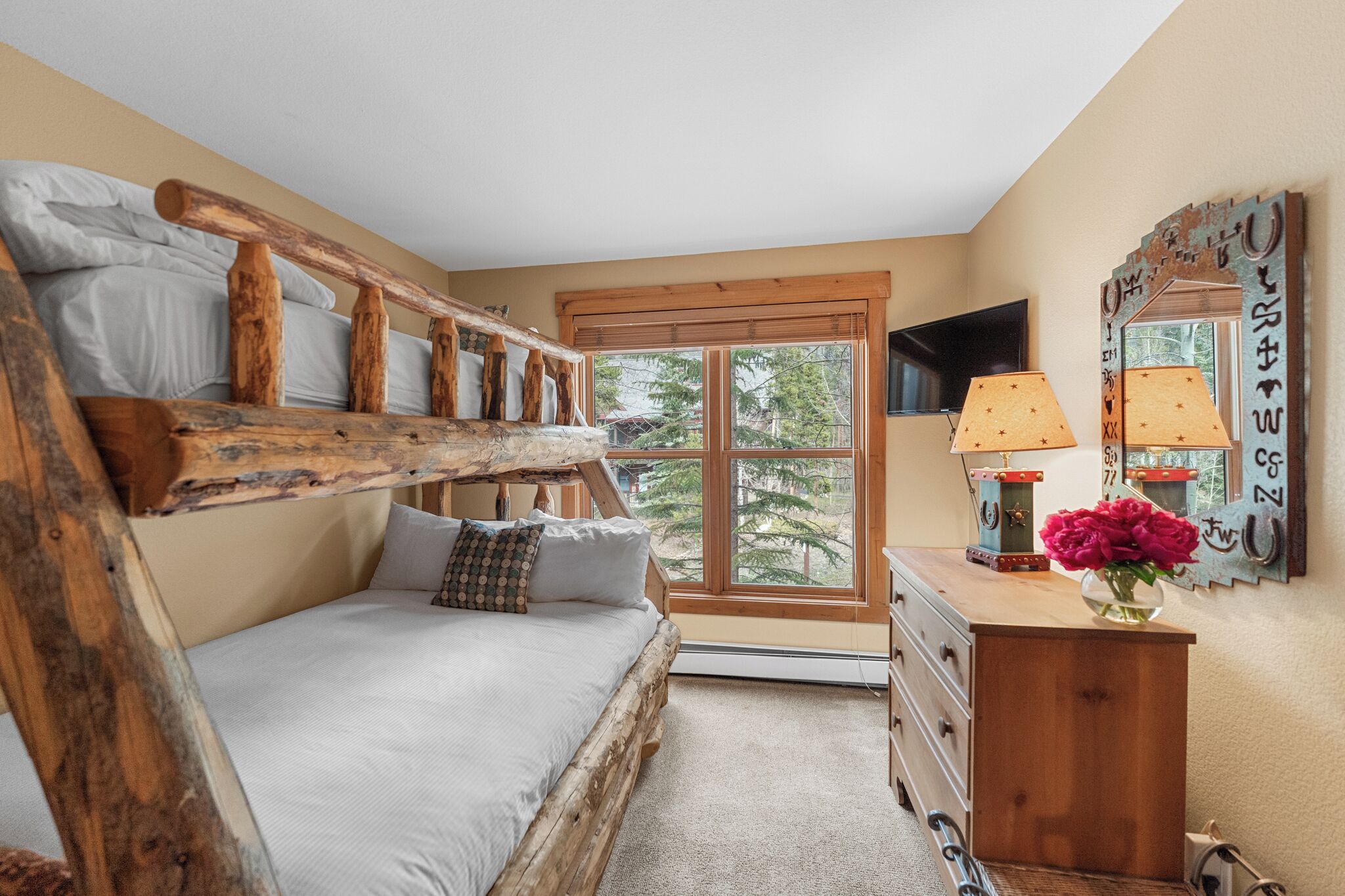 A bedroom with a bunk bed, a dresser, a mirror, and a window with a view of trees.