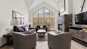 A spacious living room with a high ceiling, large windows, a stone fireplace, and a flat-screen TV.