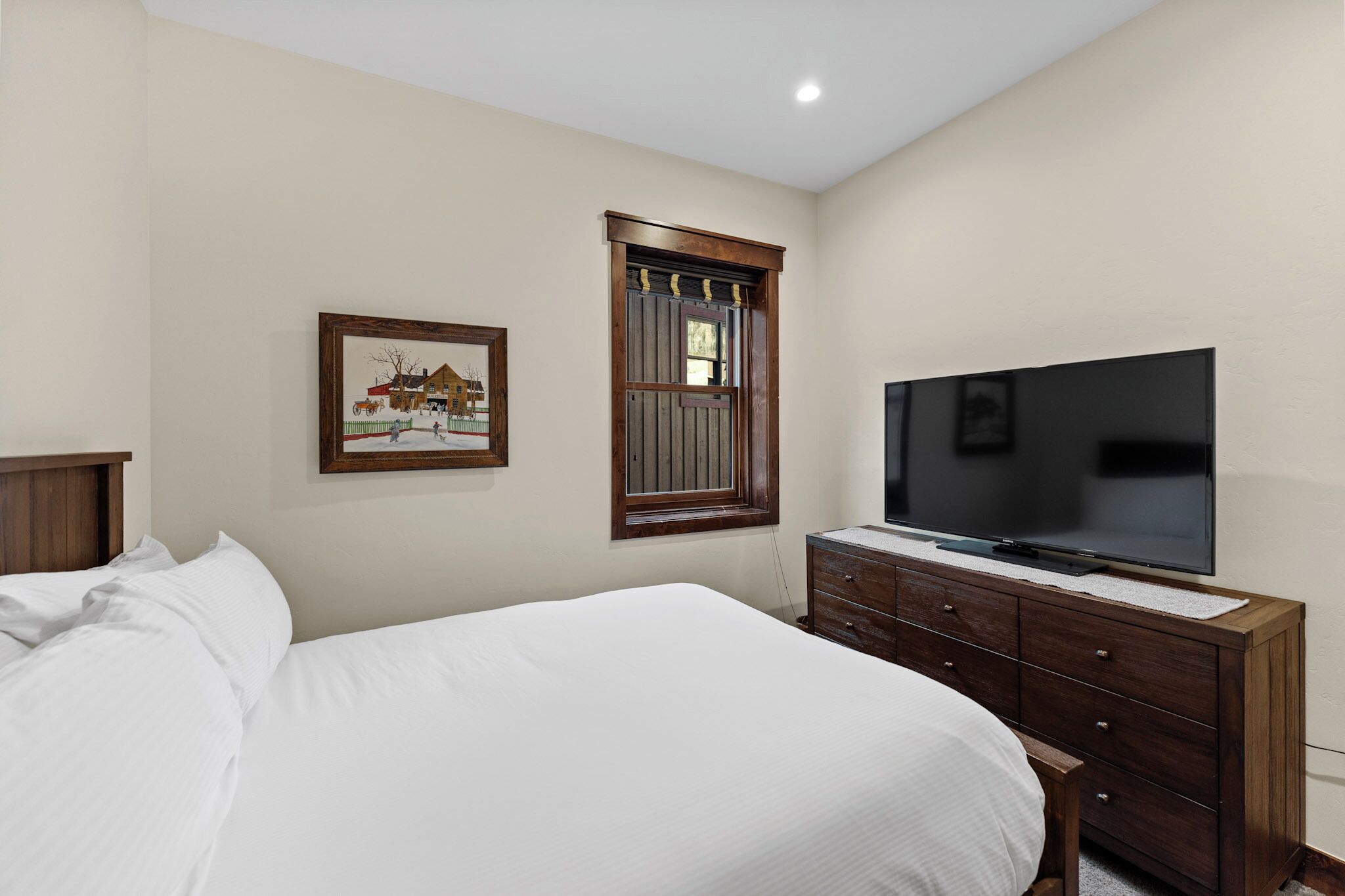 Une chambre à coucher comprenant un lit, une commode, un téléviseur, une fenêtre avec des rideaux et un tableau encadré accroché au mur.