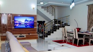 Un salon moderne doté d’un grand téléviseur à écran plat, d’un lustre, d’un canapé, d’un coin repas avec des chaises et d’un escalier.
