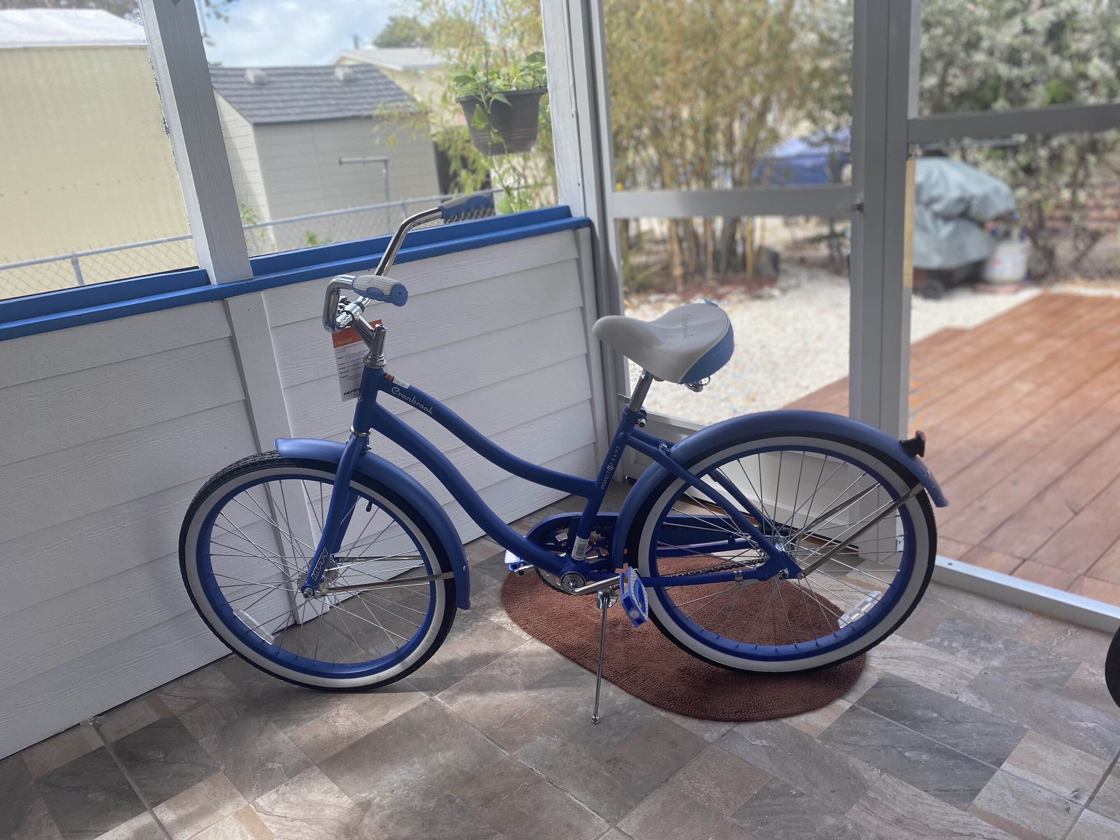 Un vélo bleu est appuyé contre un mur blanc, à l’intérieur d’une pièce d’où l’on aperçoit un jardin.