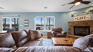 A living room with a leather sofa, a fireplace, and a view of trees through large windows. - Lakefront Oasis, Dog-Friendly with Boat and Sauna included (Pocono Summit)