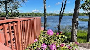 A wooden deck with a railing overlooking a lake, surrounded by trees and blooming rhododendrons. - Lakefront Oasis, Dog-Friendly with Boat and Sauna included (Pocono Summit)