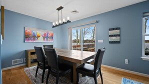 A dining area with a wooden table and chairs, a colorful painting on the wall, and a view through the window. - Lakefront Oasis, Dog-Friendly with Boat and Sauna included (Pocono Summit)