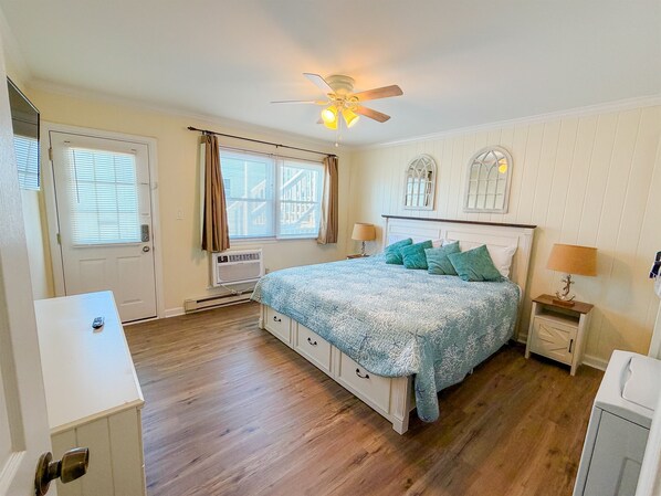 A bedroom with a bed, a ceiling fan, a window with curtains, a heater, and a nightstand with a lamp.