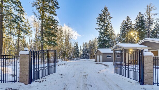A snow-covered driveway leading to a gated property with a house and a small building, surrounded by tall evergreen trees. - Columbia Falls Private Retreat: Pool Table & Deck! (Columbia Falls)