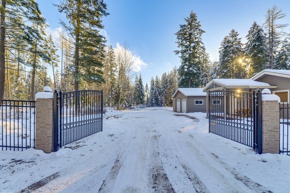 A snow-covered driveway leading to a gated property with a house and a small building, surrounded by tall evergreen trees.