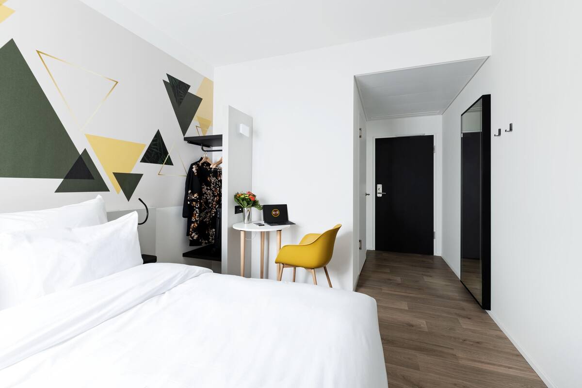 Habitación de hotel moderna con una cama bien hecha, un escritorio pequeño con una computadora portátil, una silla amarilla y un diseño geométrico en la pared.