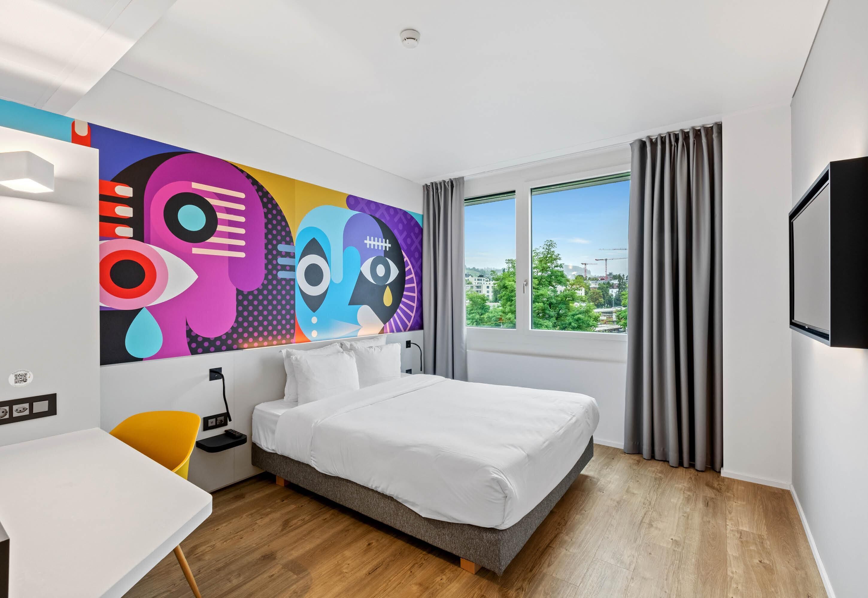 Una camera d'hotel moderna con un grande letto, una scrivania, un murale astratto dai colori vivaci e vista sulla città.
