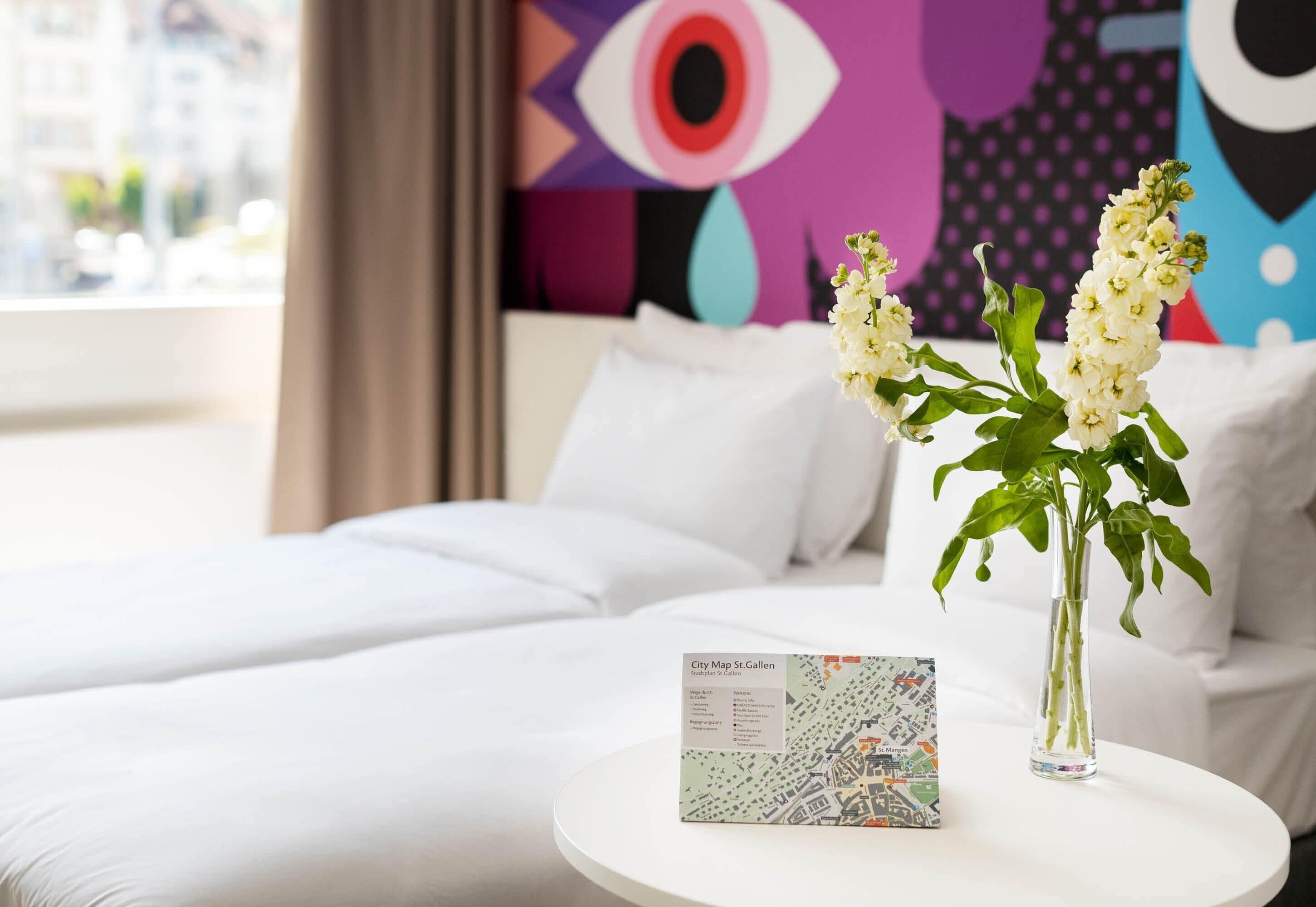 Een hotelkamer met een bed, een witte ronde tafel met een kaart, en een vaas met bloemen.