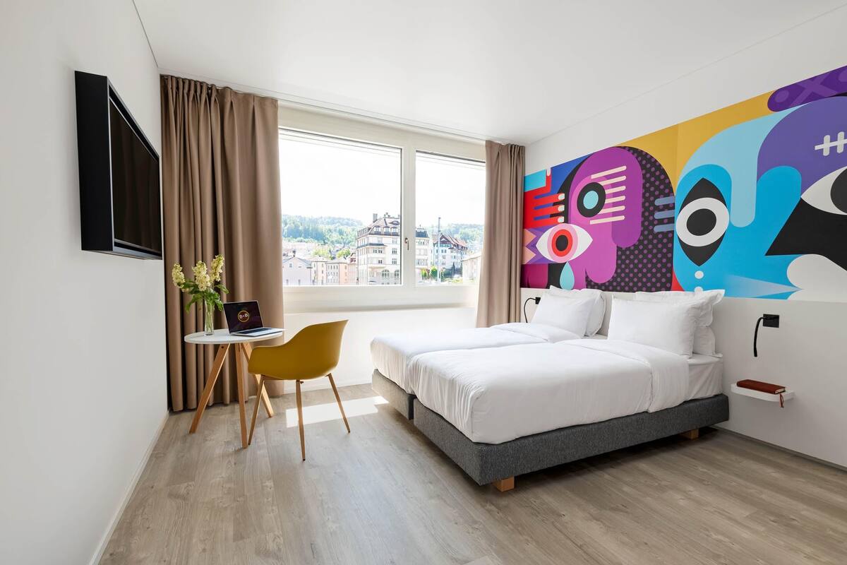 A hotel room with a bed, a desk with a laptop, a chair, a window with curtains, and a colorful abstract painting on the wall.