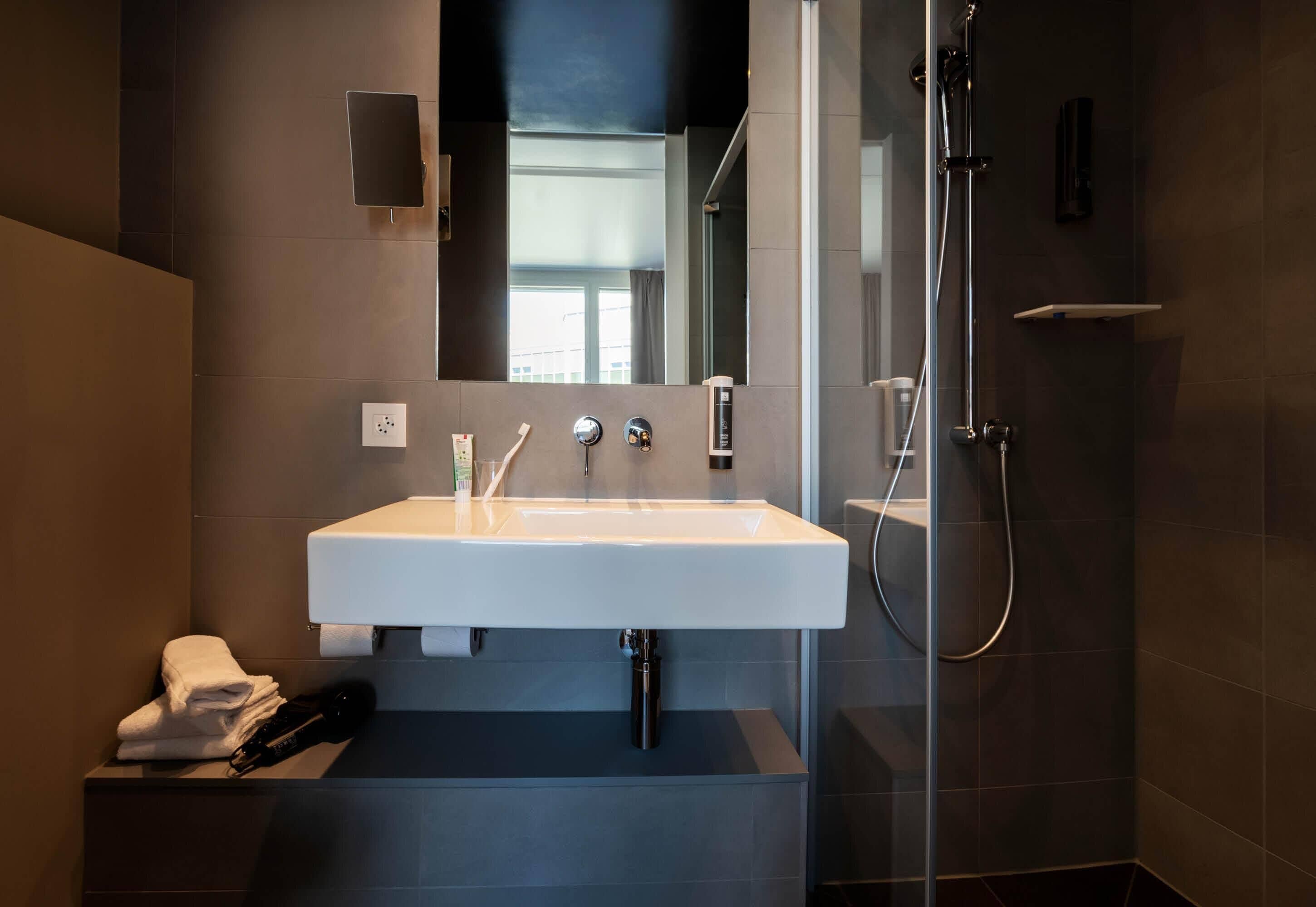 Ein modernes Badezimmer mit einem weißen Waschbecken, einem Spiegel, einer Dusche und einem Handtuchhalter.