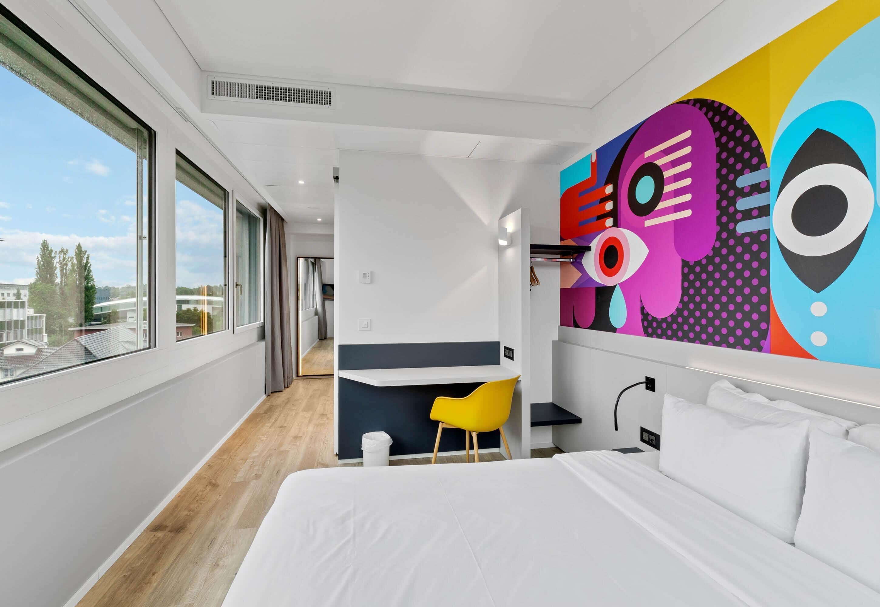 Una habitación de hotel moderna con una cama grande, un escritorio, una silla amarilla y una obra de arte abstracta y colorida en la pared.
