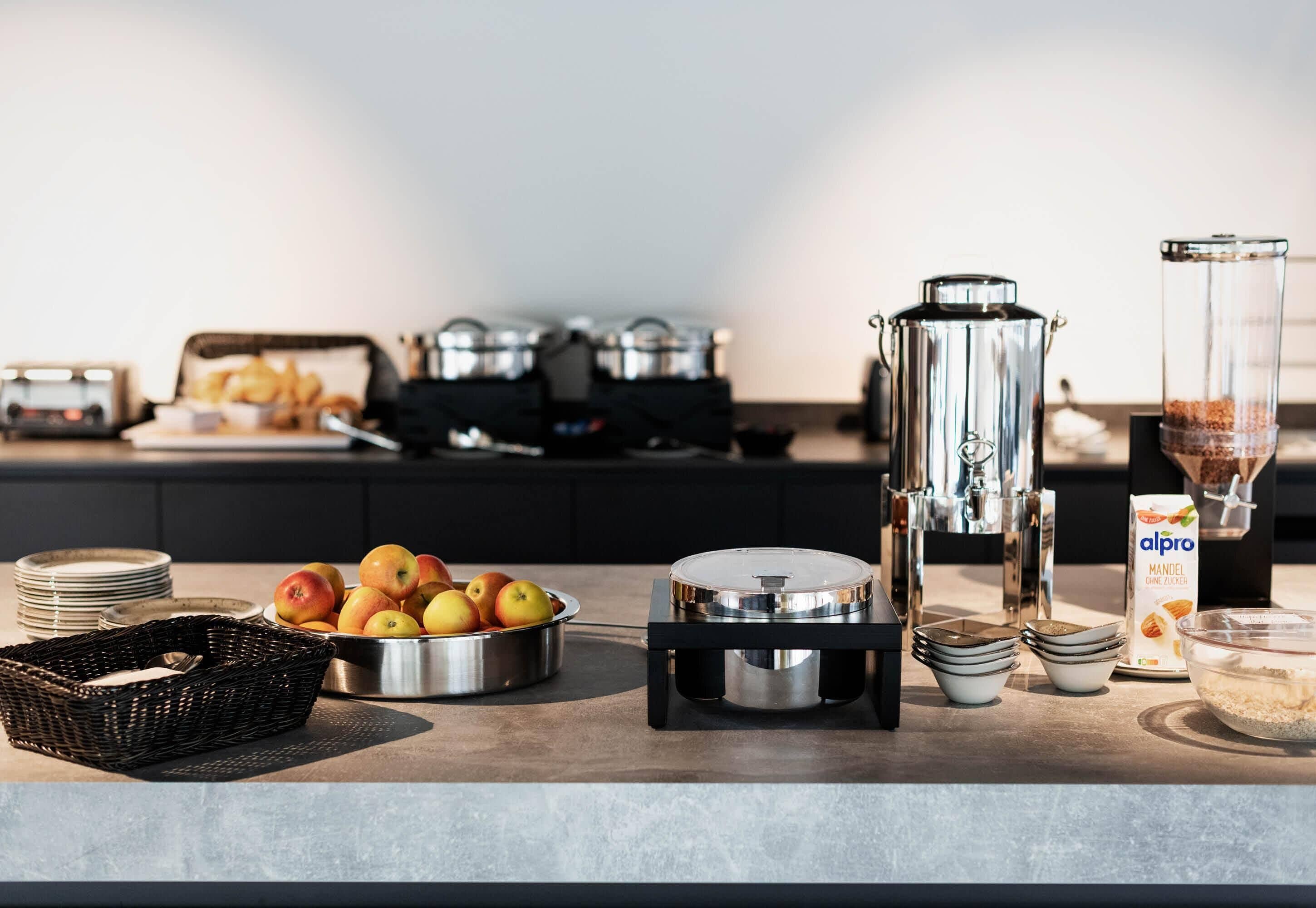 Mesa de desayuno buffet con cafetera, un bol de manzanas y un cartón de leche de almendras.