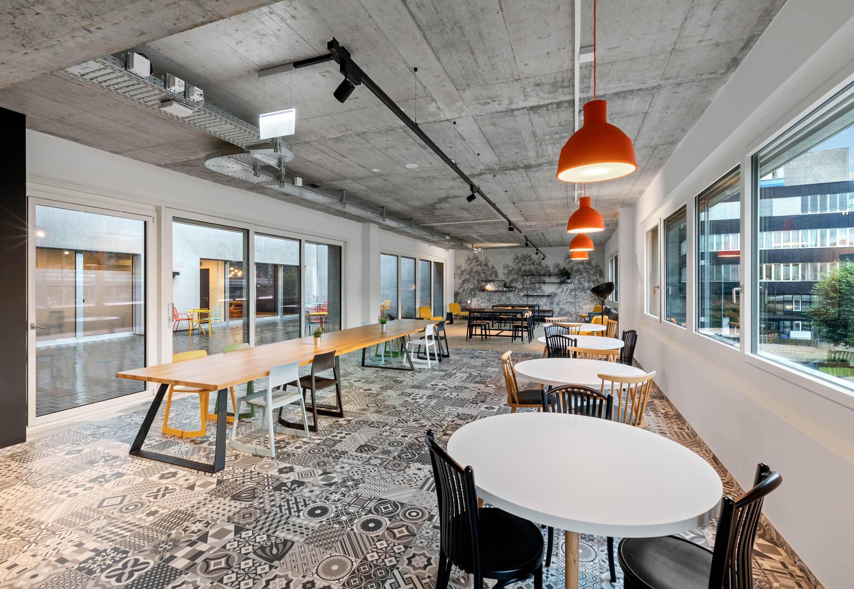A modern office space with large windows, patterned carpet, and a mix of round and rectangular tables.