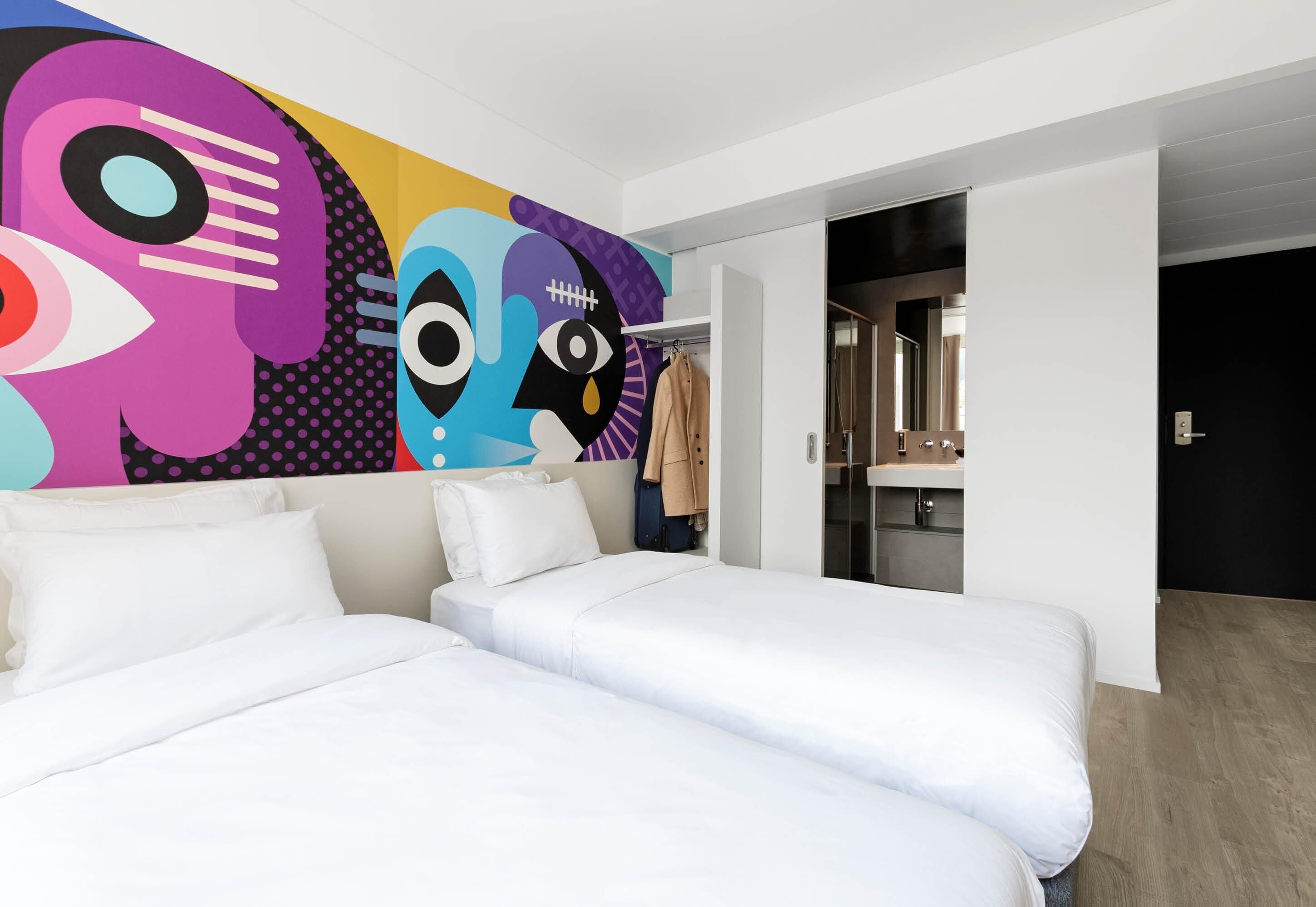 A hotel room with two beds, a colorful abstract mural, a coat rack, and a bathroom visible through an open door.