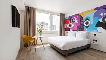 A modern hotel room with a bed, a small table with a plant, a yellow chair, and a large abstract painting on the wall.