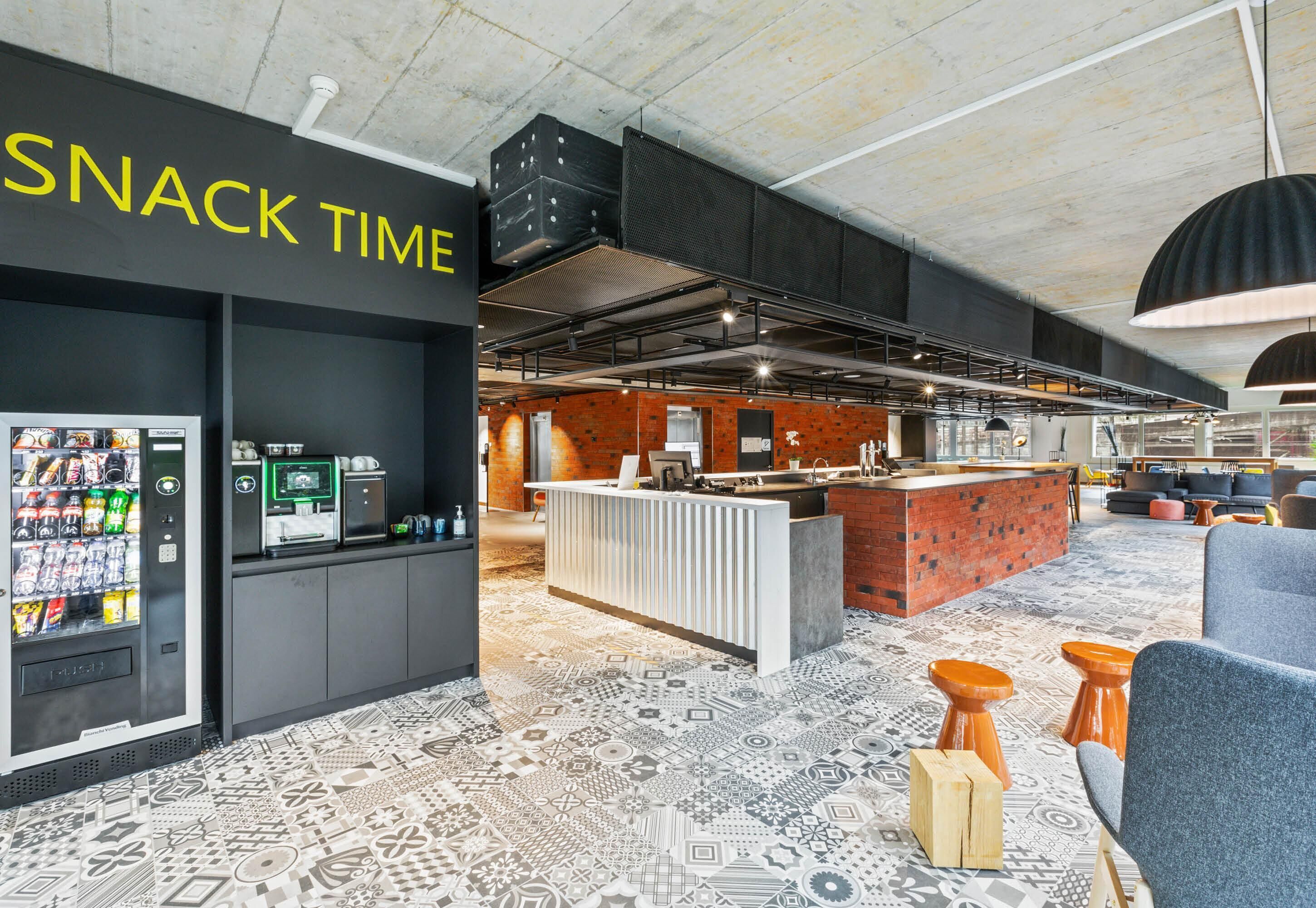 A modern lounge area with a vending machine labeled 'SNACK TIME,' a bar counter, and patterned flooring.