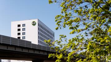 A modern building with a B&B logo, a bridge in the foreground, and green foliage in the background.