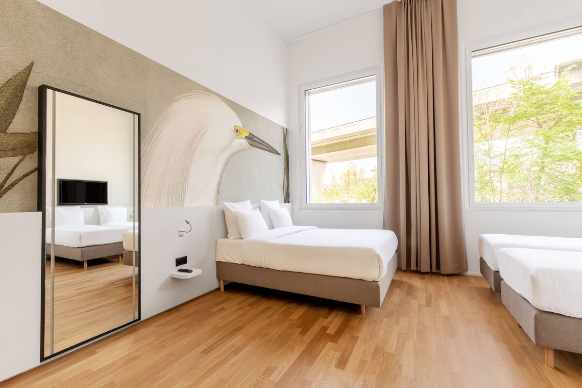 A modern hotel room with a large bed, a smaller bed, a mirror, and a window with curtains.