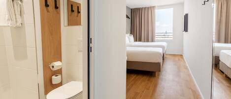 Habitación de hotel con ducha, inodoro, dos camas y un ventanal grande con cortinas.