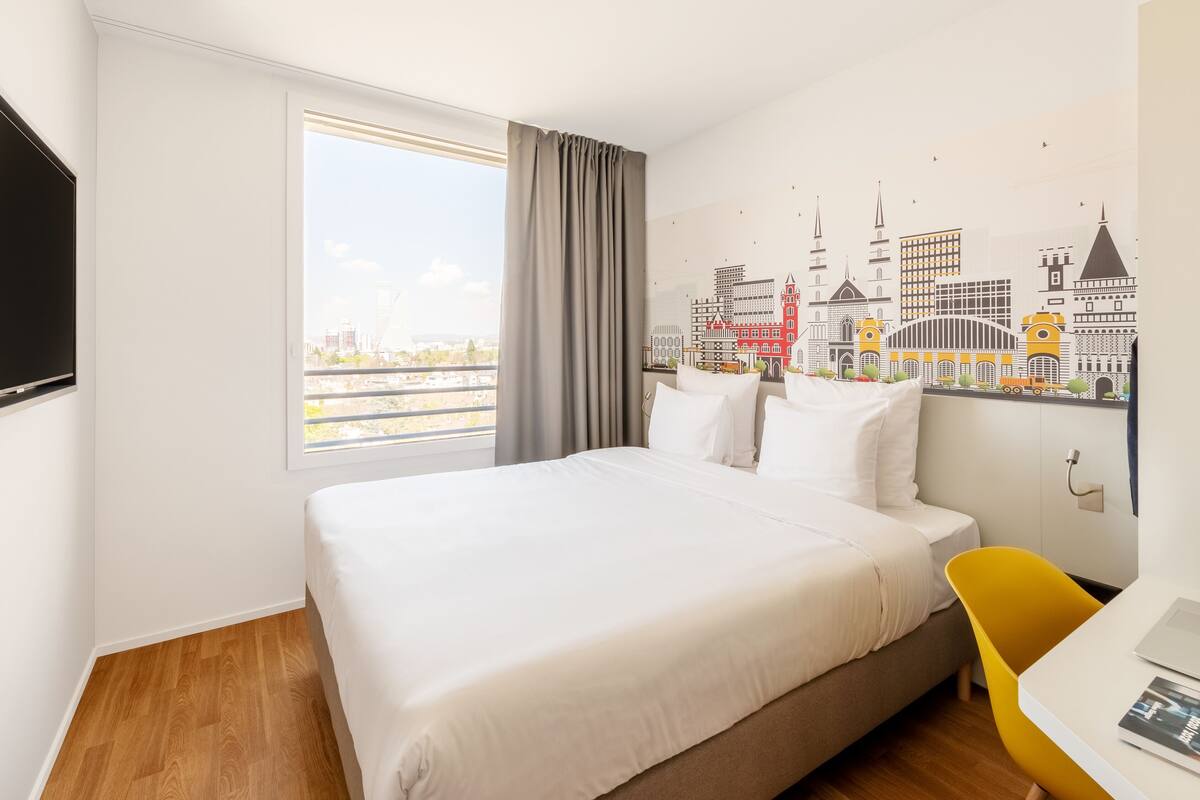 A hotel room with a bed, a desk, a chair, a TV, and a cityscape mural on the wall.