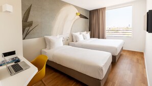 Habitación de hotel con dos camas, un escritorio con una computadora portátil, una silla y un amplio ventanal con vista a la ciudad.