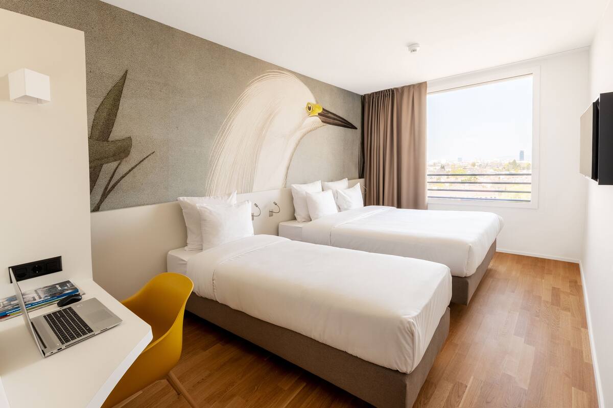 A hotel room with two beds, a desk with a laptop, a chair, and a large window with a view of the city.