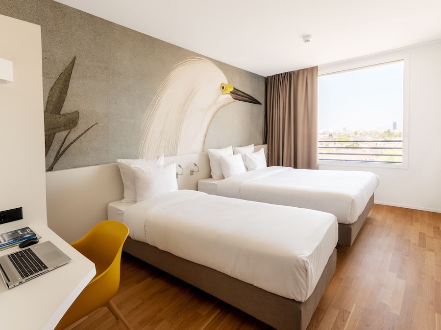 Habitación de hotel con dos camas, un escritorio con una computadora portátil, una silla y un amplio ventanal con vista a la ciudad.