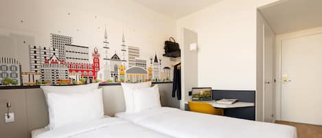 Ein Hotelzimmer mit Bett, Schreibtisch, Stuhl, Kleiderhaken und Wandbild mit Stadtansichten.