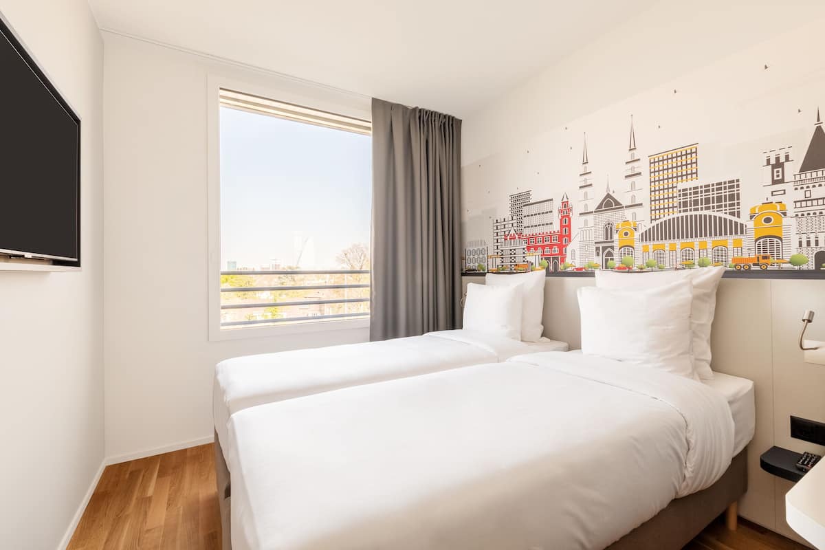A hotel room with two beds, a large window, and a cityscape mural on the wall.
