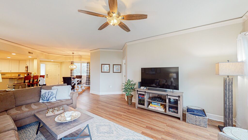 A spacious living room with a large flat-screen TV, a ceiling fan, and a dining area in the background.