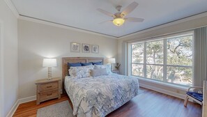 A bedroom with a large bed, two bedside tables, a ceiling fan, and a large window with a view of trees. - Large Two Bedroom Villa with Ocean Views! (Hilton Head)