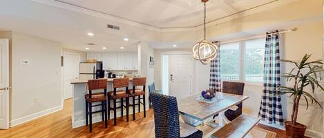 A spacious dining area with a wooden table and chairs, a hanging light fixture, and a view of the kitchen in the background.