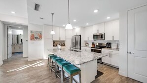 Cocina moderna con armarios blancos, una isla de granito con taburetes de bar, electrodomésticos de acero inoxidable e iluminación colgante.