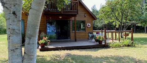 Una cabaña de madera con balcón, rodeada de árboles y un patio cubierto de césped.